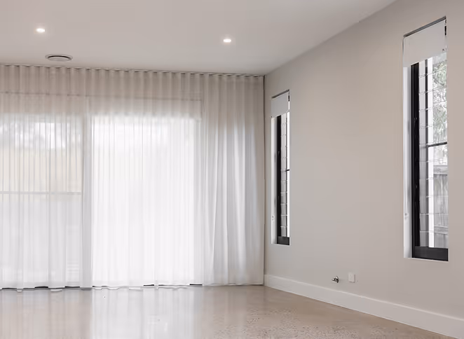Empty room with polished floor, sheer white curtains on large window, and two tall narrow windows with blinds.