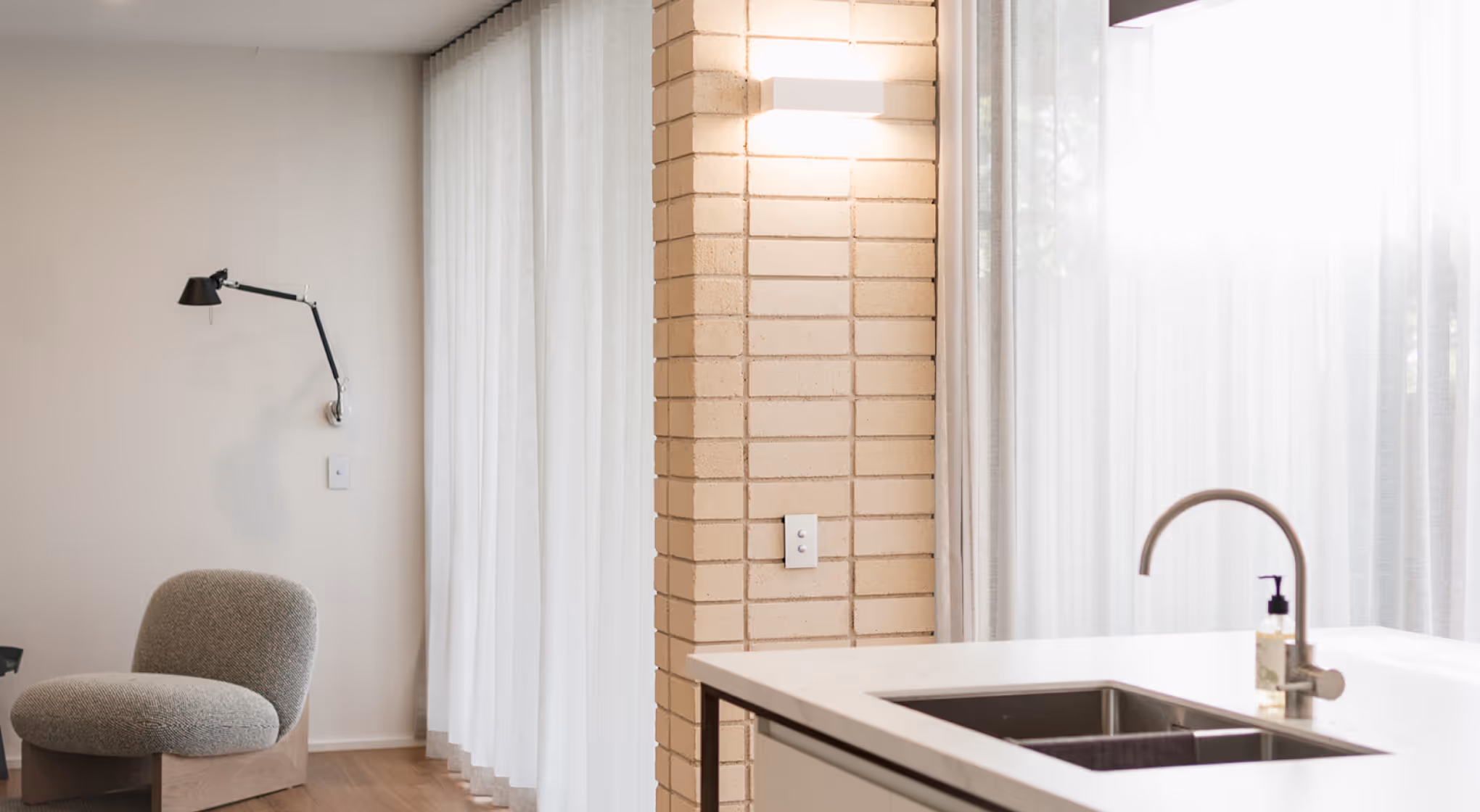 Modern kitchen sink with curved faucet next to a brick column and a gray cushioned chair near a wall lamp.