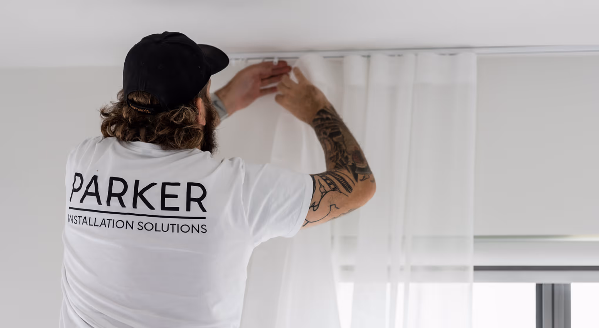 Man with tattoos wearing a Parker Installation Solutions shirt hangs white curtains on a rail.