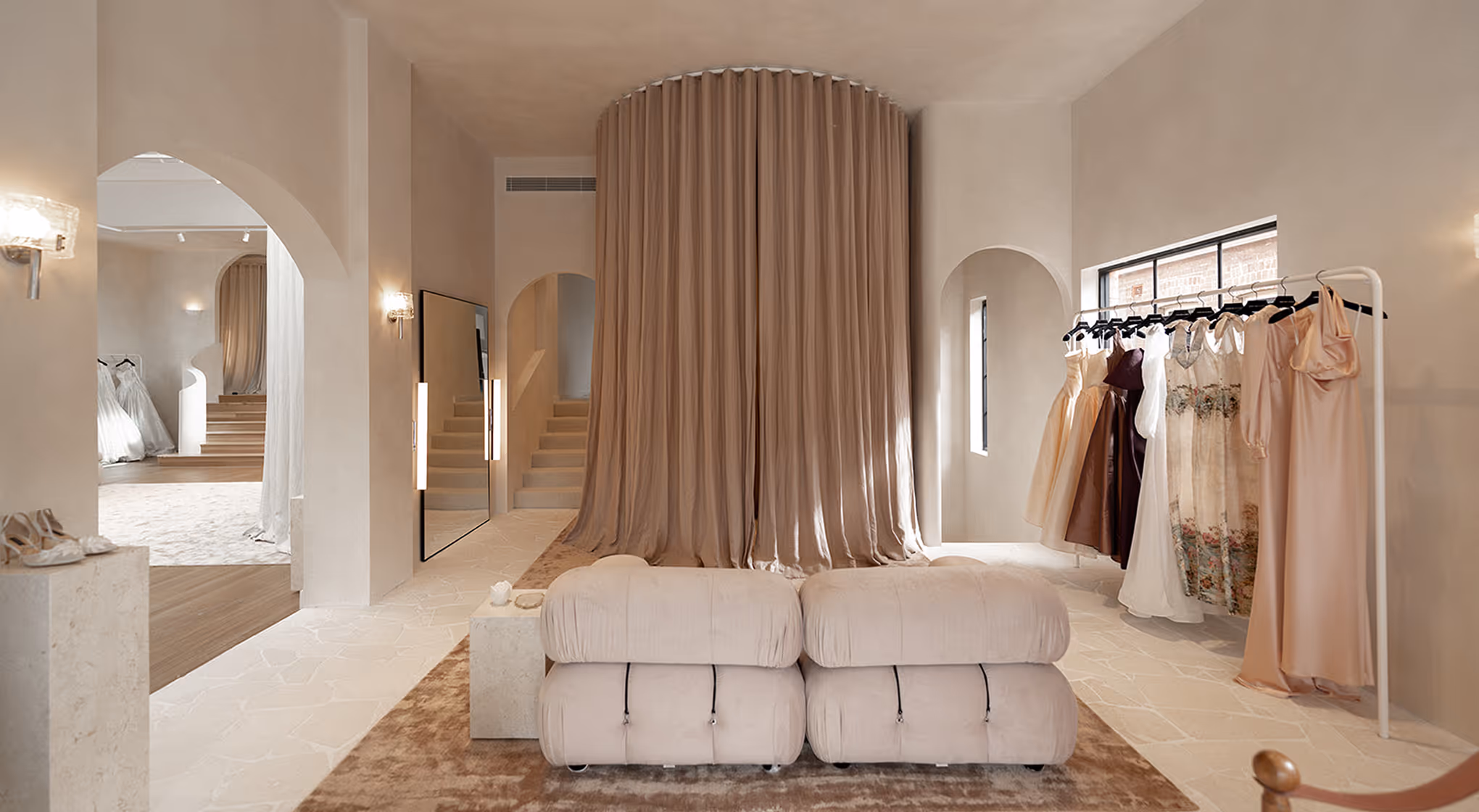 Elegant boutique interior with soft beige tones, dresses on rack, plush seating, and flowing curtain.