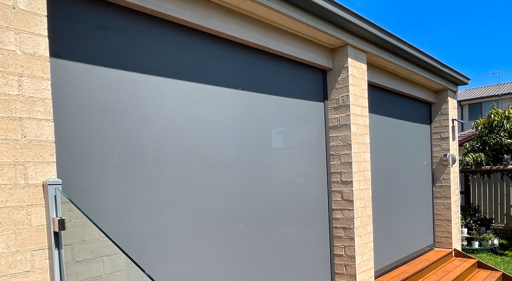 Two large gray outdoor roller shades on a brick house patio under clear blue sky.