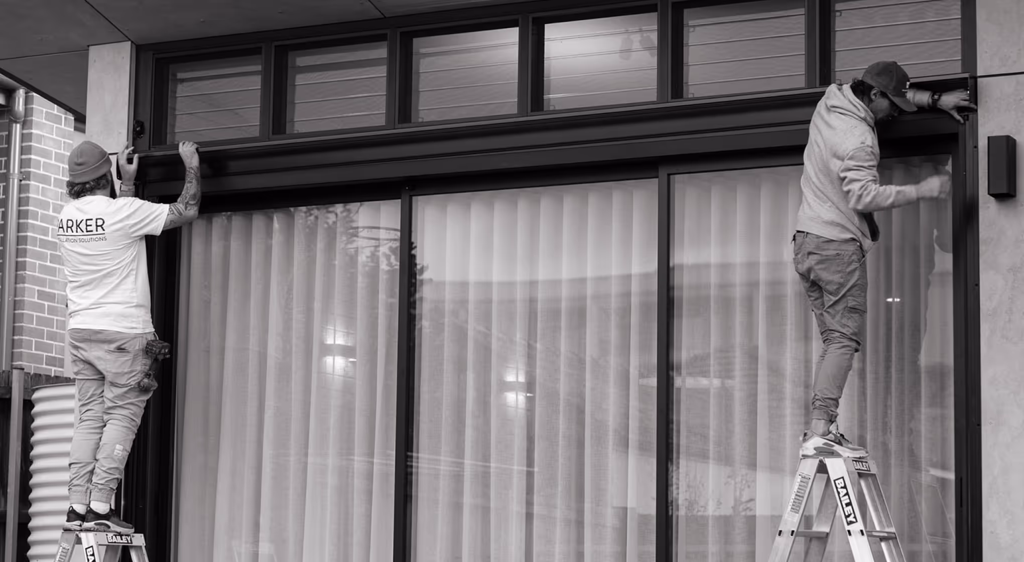 Two workers on ladders installing or fixing a black frame on a large window with vertical blinds.