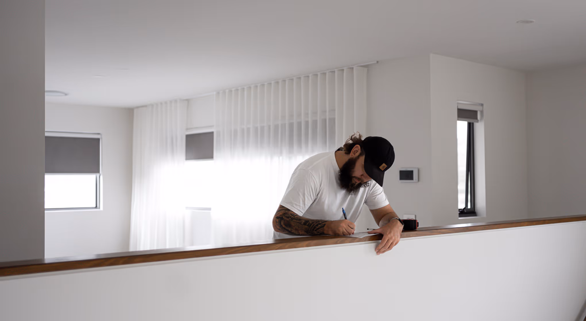 Bearded man in a black cap writing on paper while leaning on a wooden railing indoors.