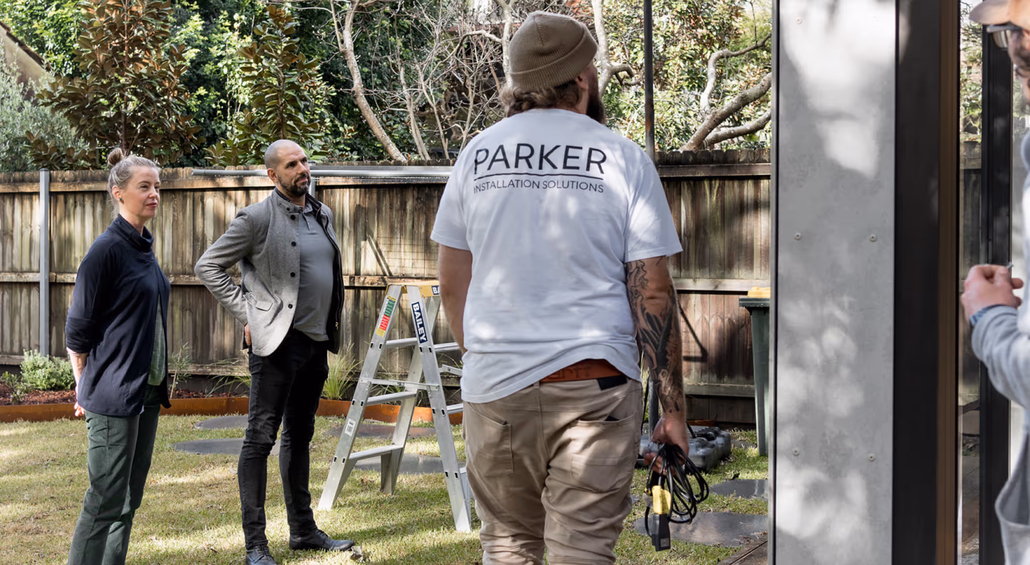 Man in white Parker Installation Solutions shirt holds cables, two people watch outside near ladder.
