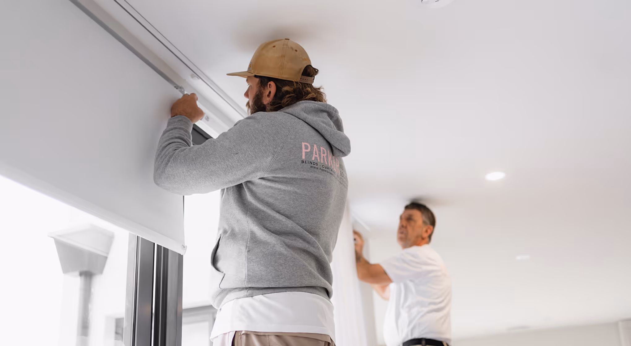 Two men installing white roller blinds on windows in a bright room.