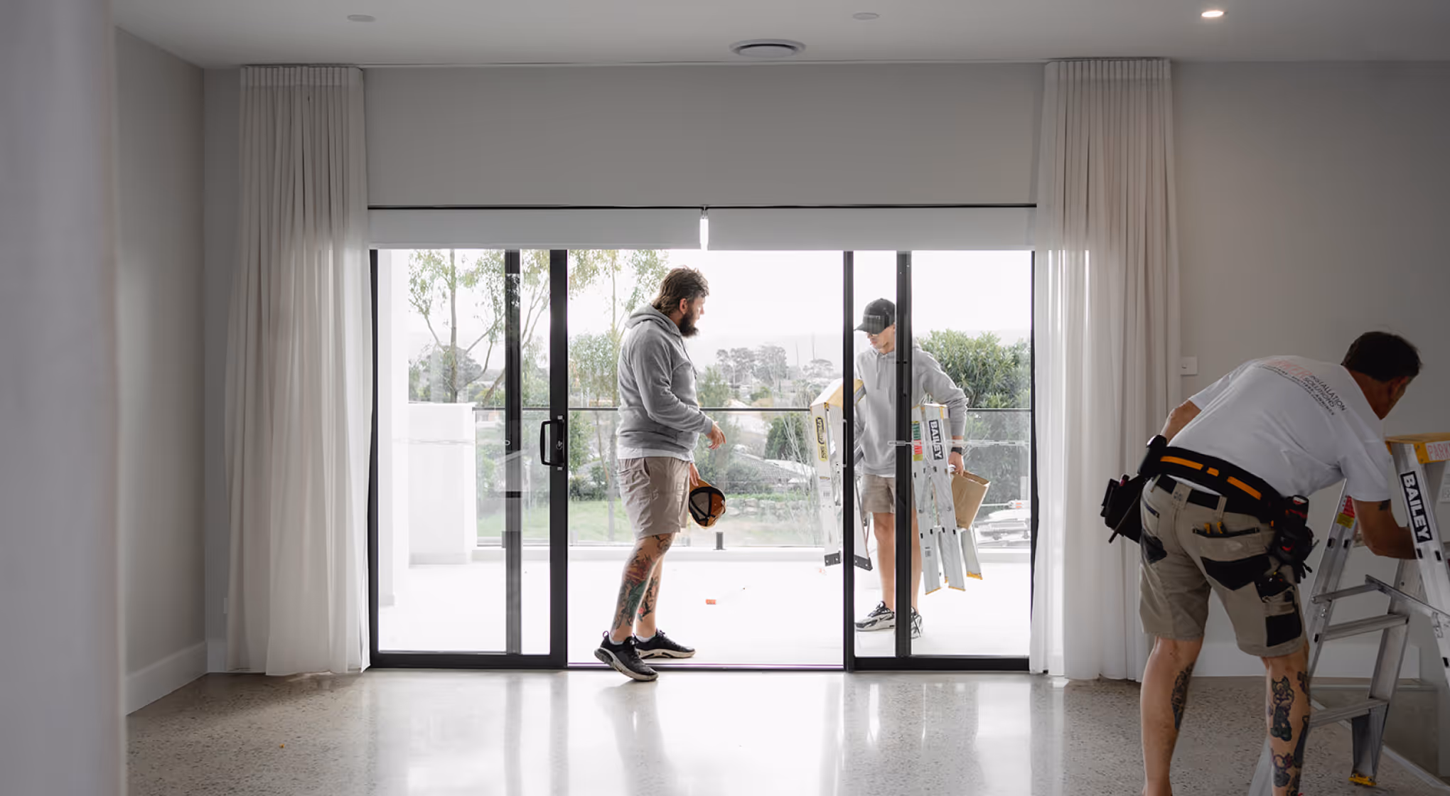 Three men with tattoos working near sliding glass doors, one holding a ladder and another adjusting it.