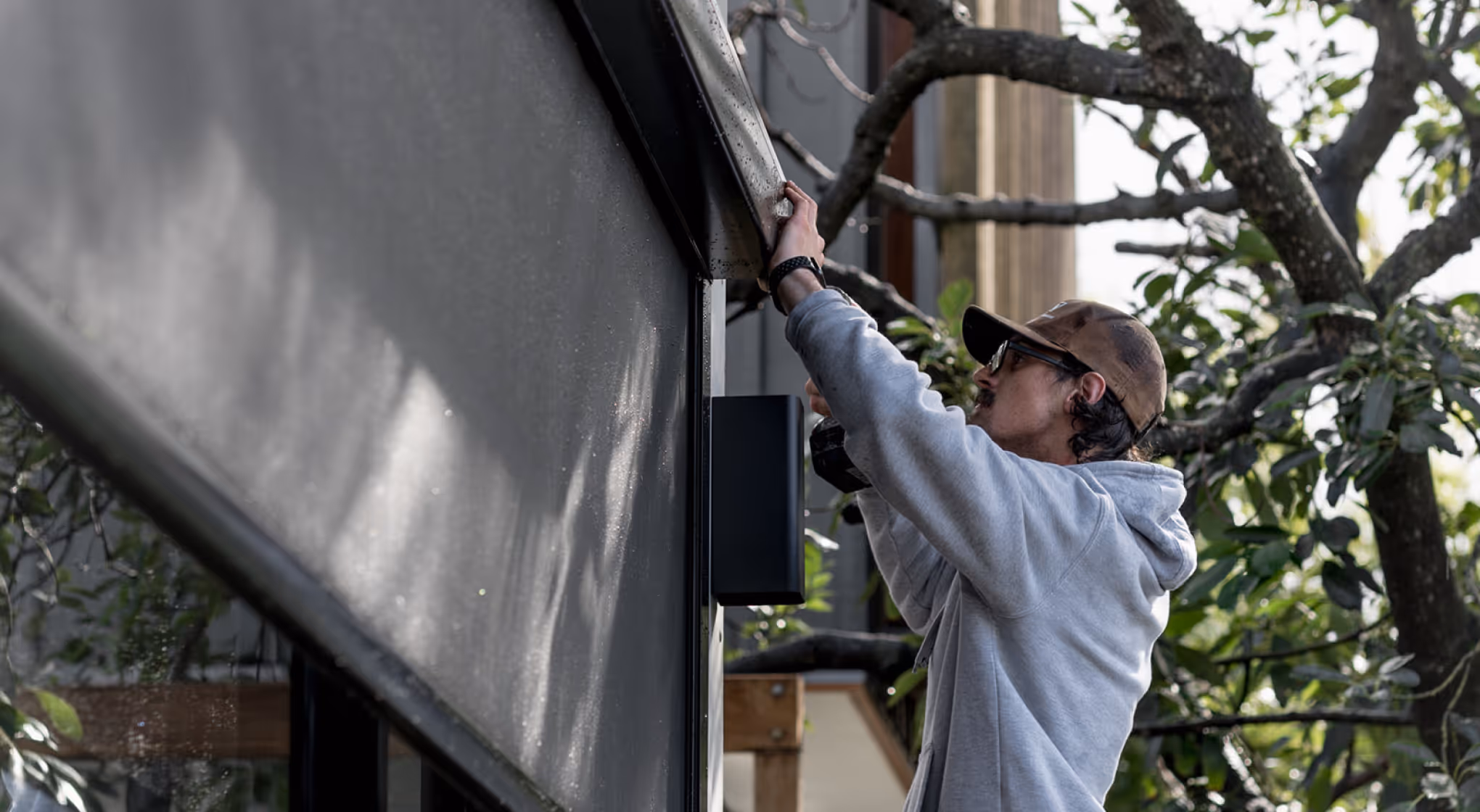 Man in cap and hoodie adjusting or installing an outdoor window shade on a building.