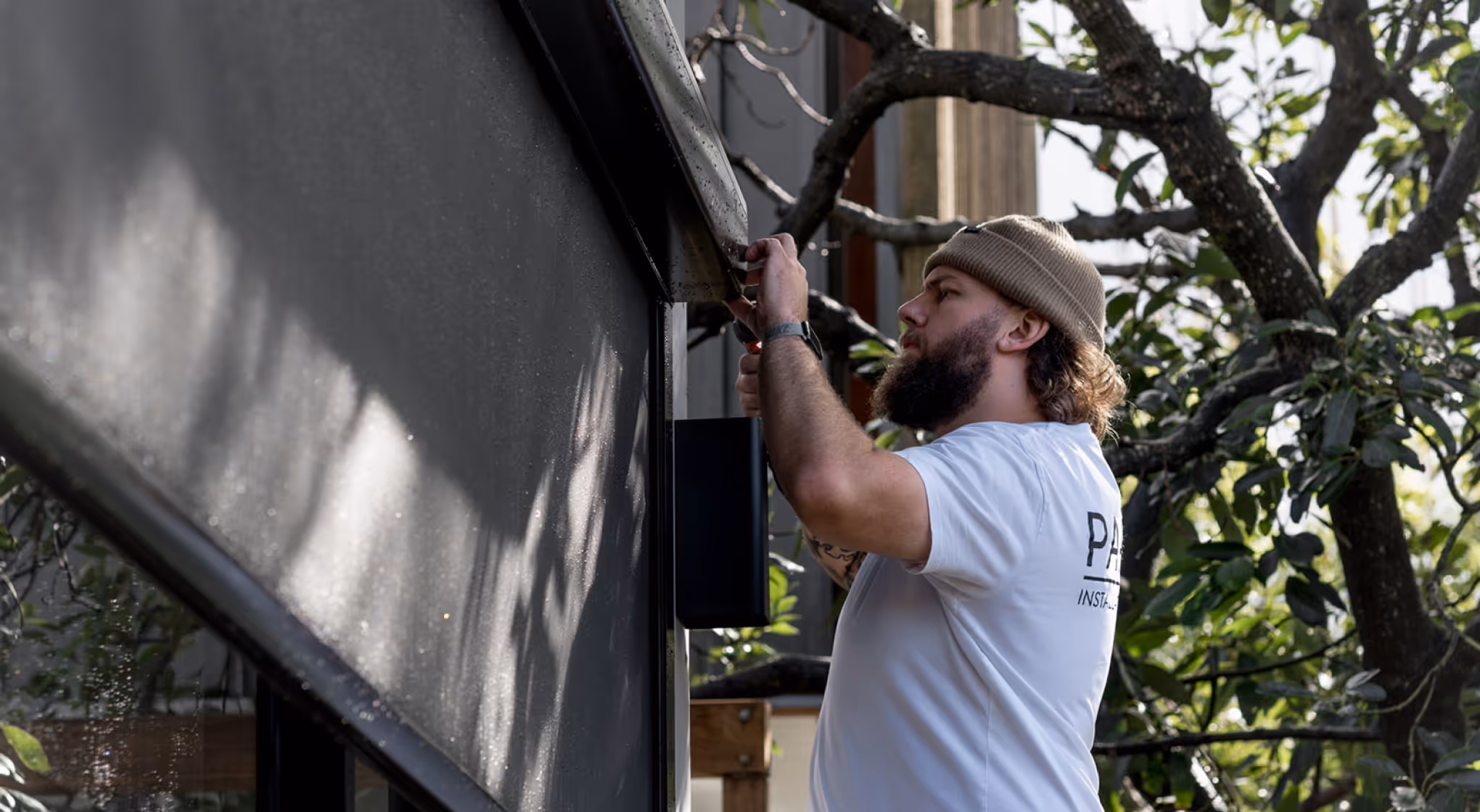 Bearded man in a beanie installs or repairs a dark outdoor fixture on a building wall.