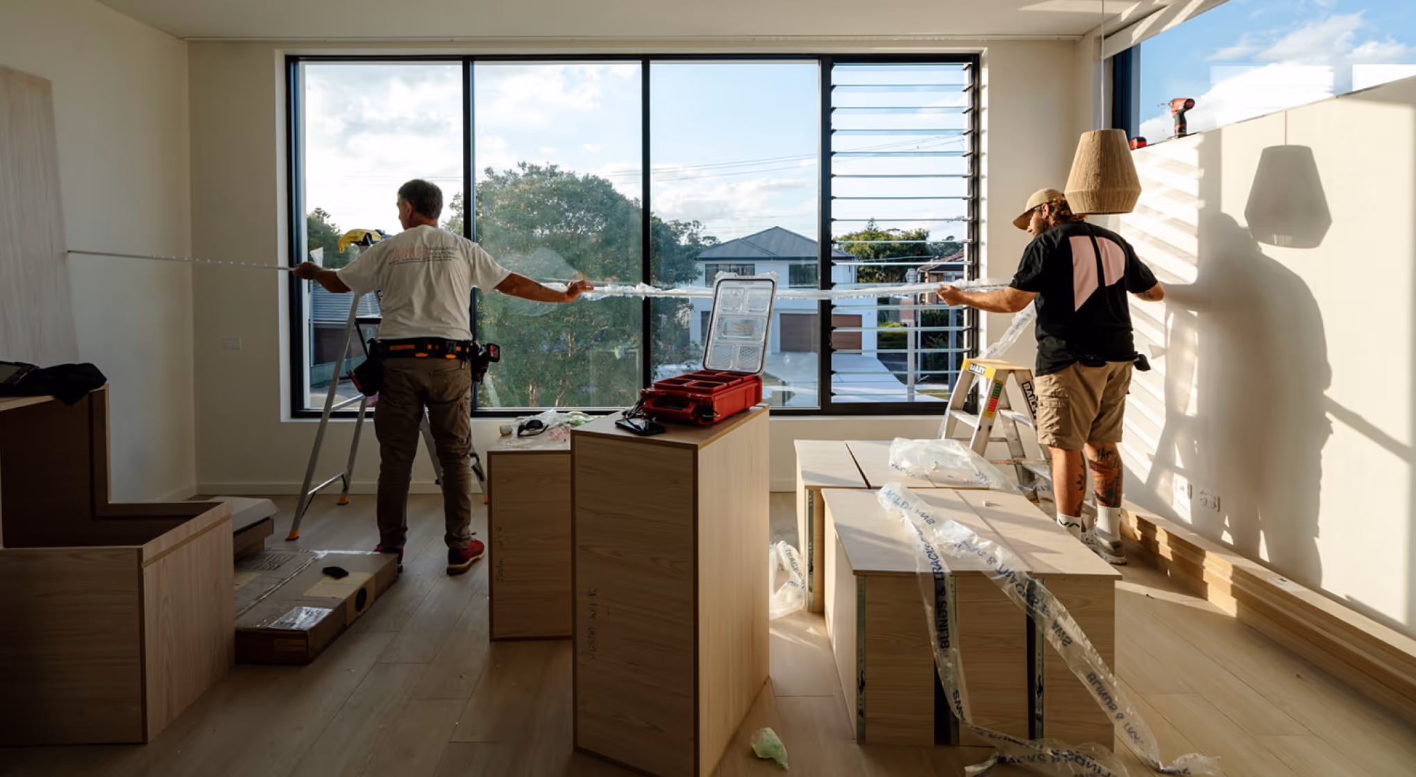 Two workers install window blinds in a sunlit room with light wood flooring and furniture.