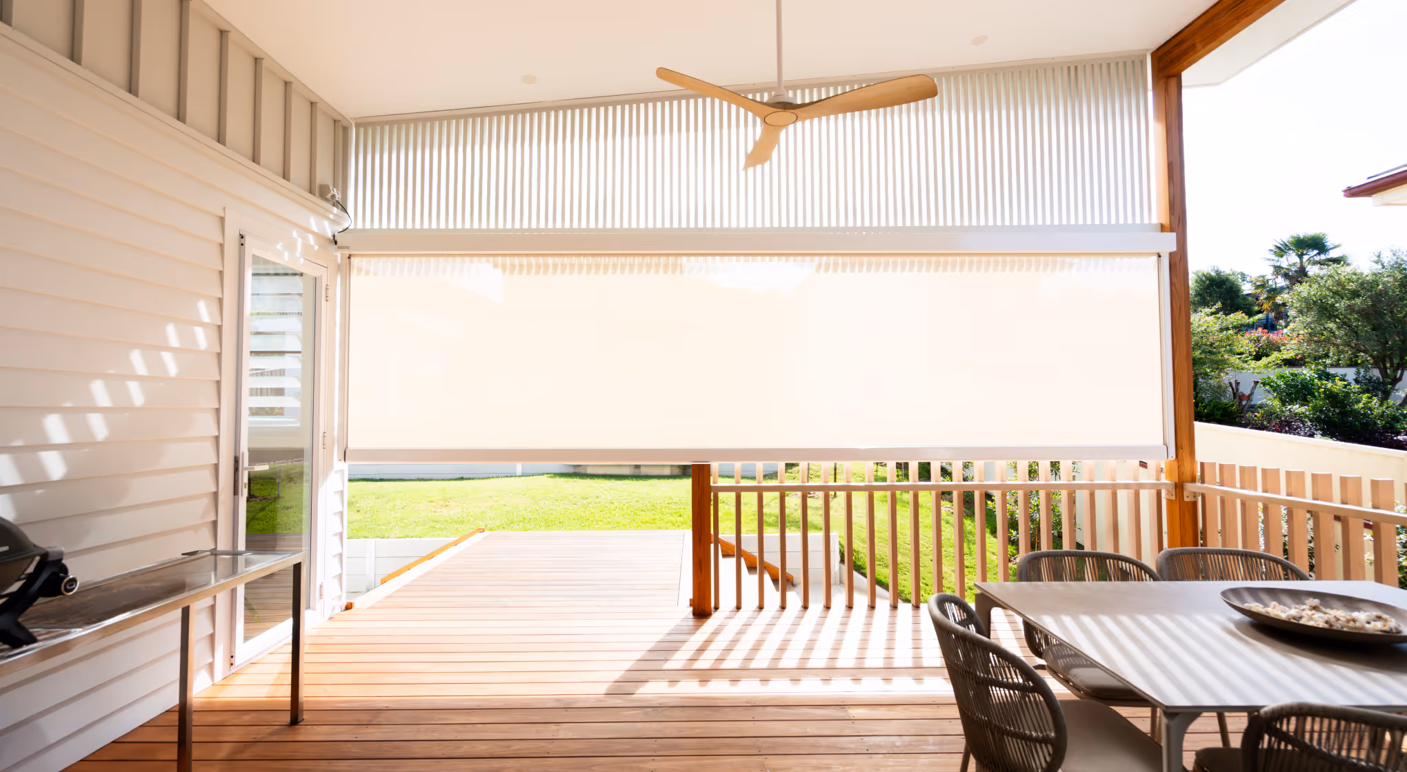 Bright patio with wooden deck, hanging fan, white retractable shade, table, and chairs overlooking green lawn.