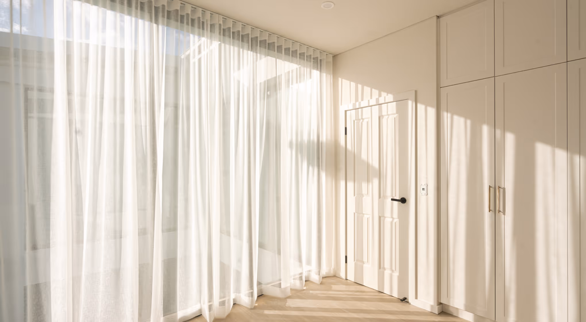 Room corner with sheer white curtains, white door, and built-in white cabinets with gold handles.