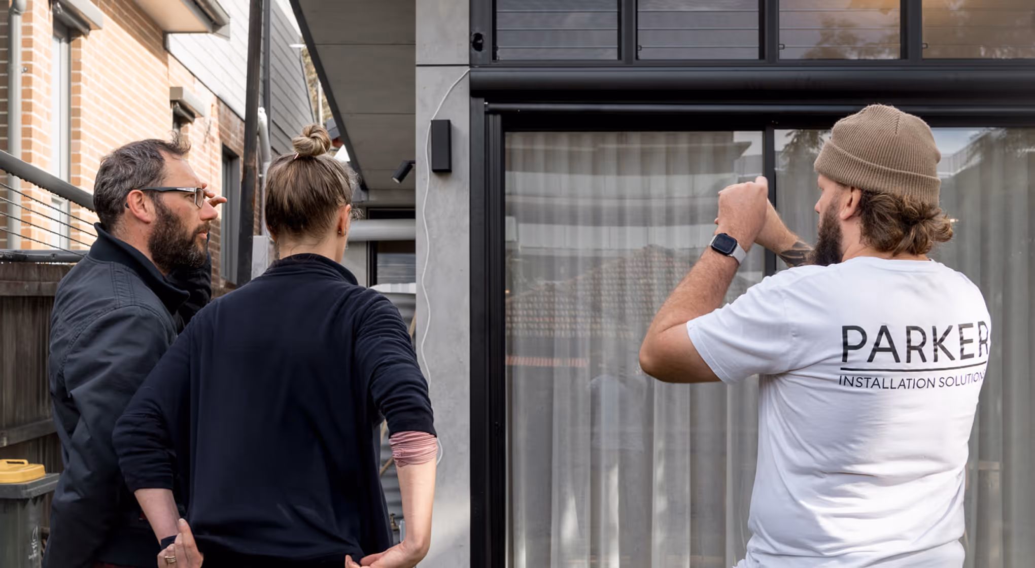 Man in white Parker shirt showing a detail to two people outside a modern building with glass doors.