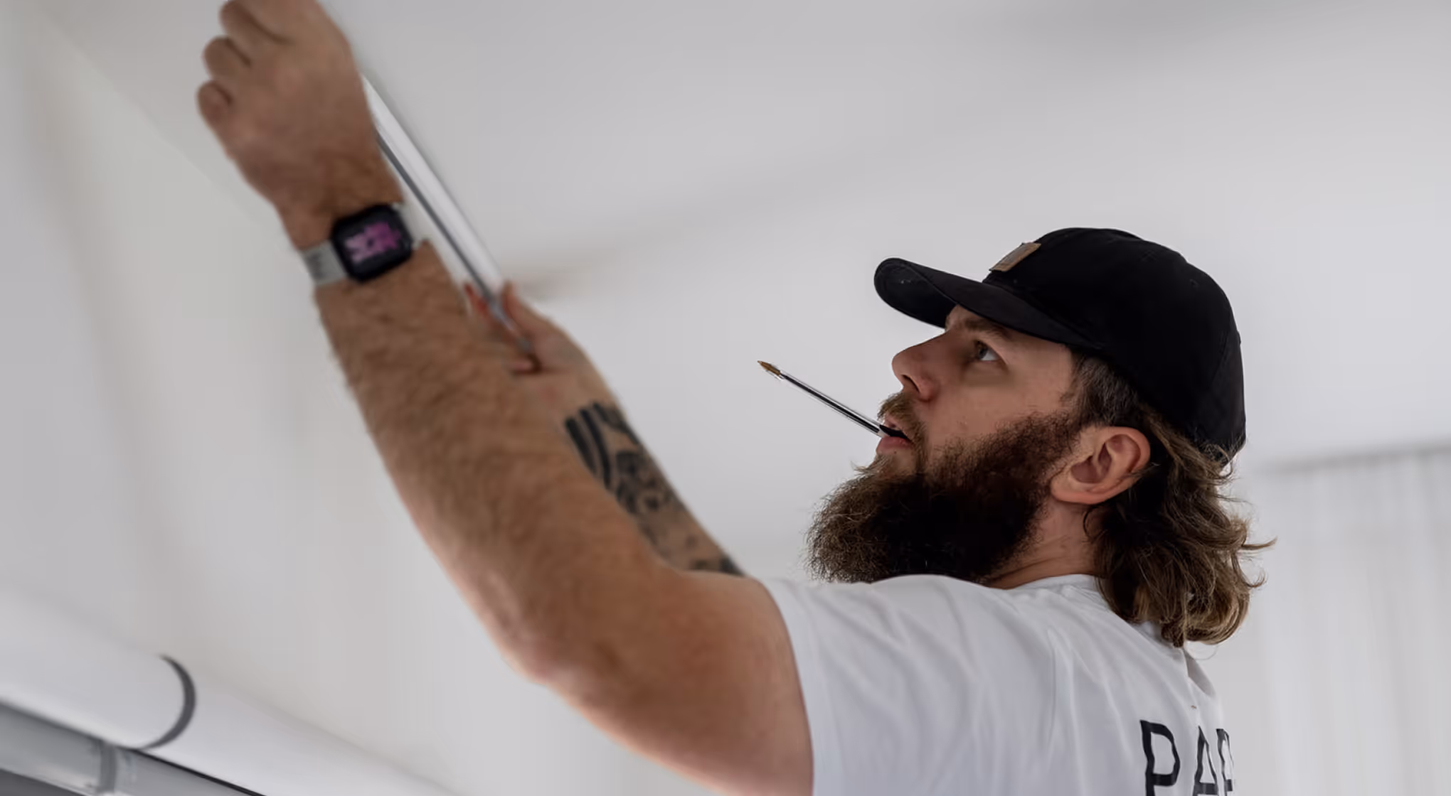 Bearded man wearing black cap and watch measuring ceiling with tape and holding pencil in mouth.