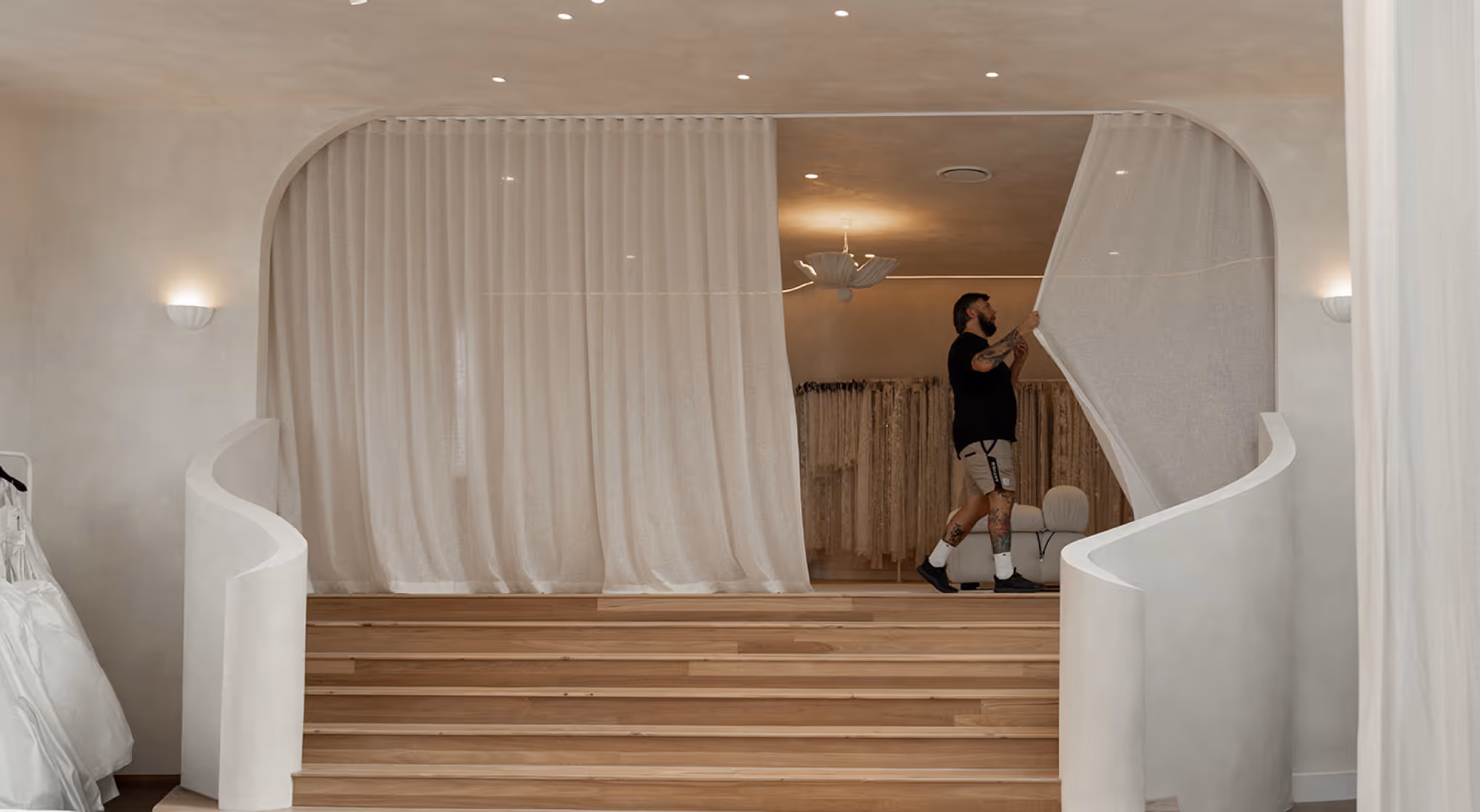 Man pulling sheer white curtain in modern room with wooden stairs and wedding dresses.