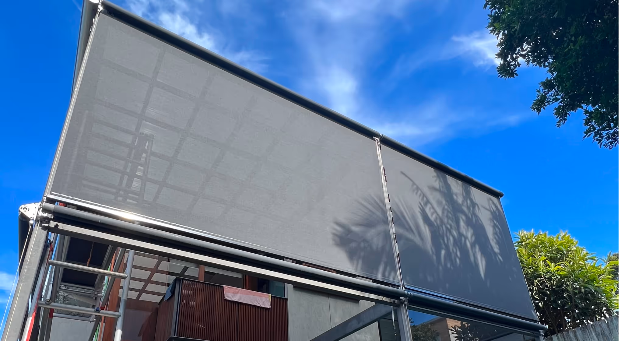 Outdoor roller shade with shadow of palm leaves on a sunny day under a blue sky.