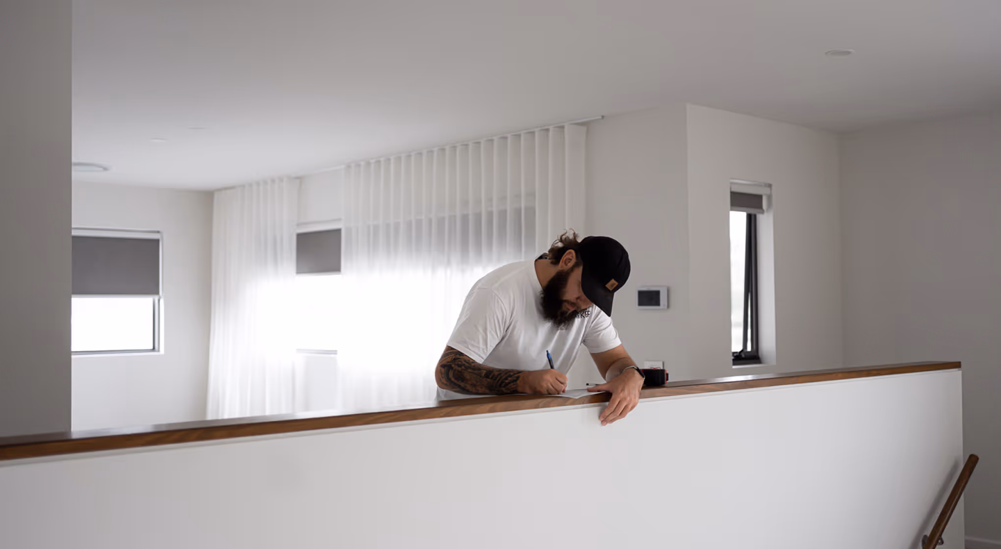 Bearded man wearing a cap writing on paper while leaning on a wooden railing indoors.