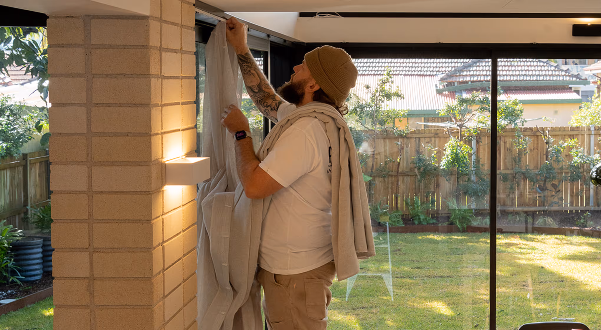 Bearded man with a beanie hanging beige curtains on a window in a sunlit room overlooking a garden.