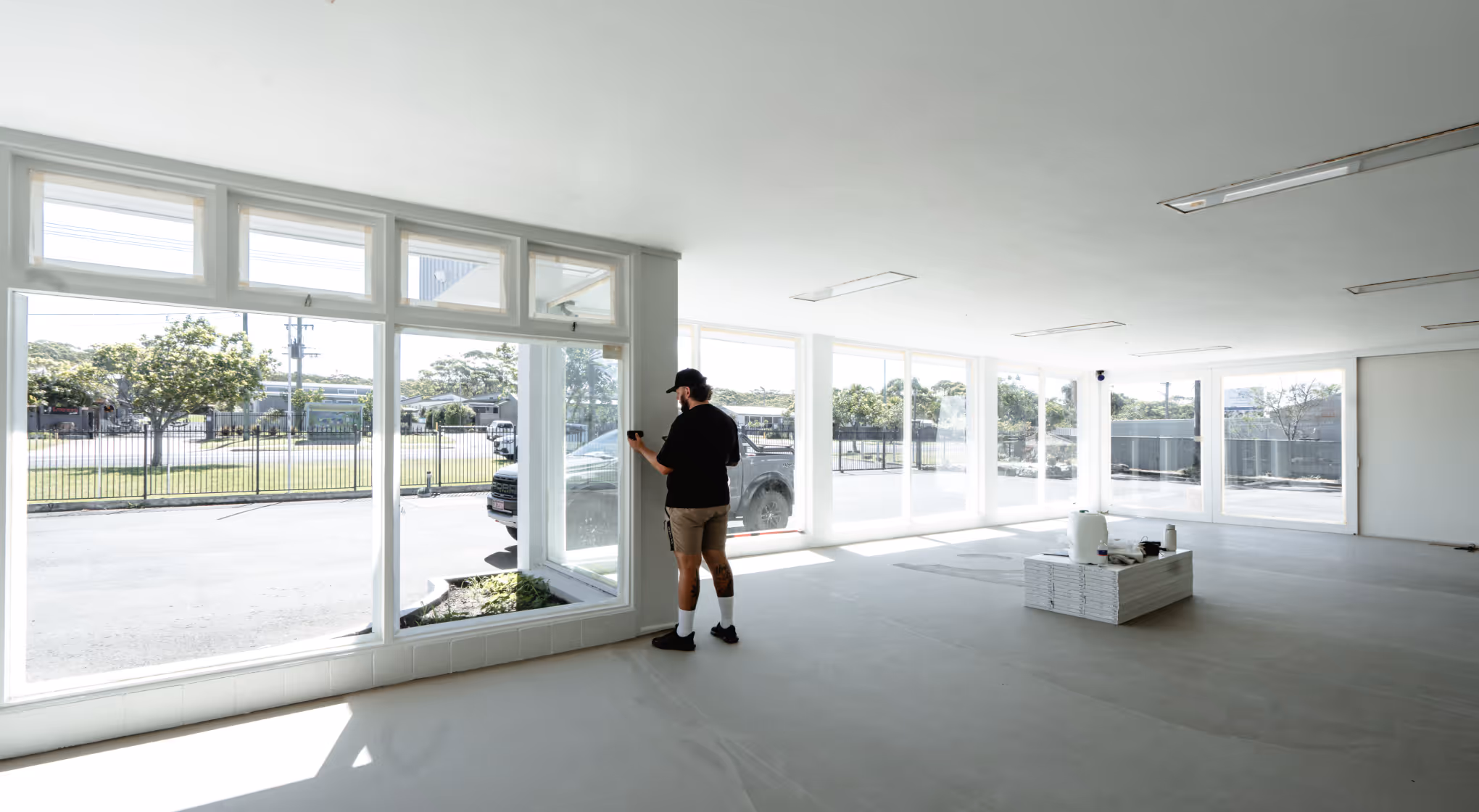 Man standing inside a bright, empty room with large windows and a small stack of tiles on the floor.