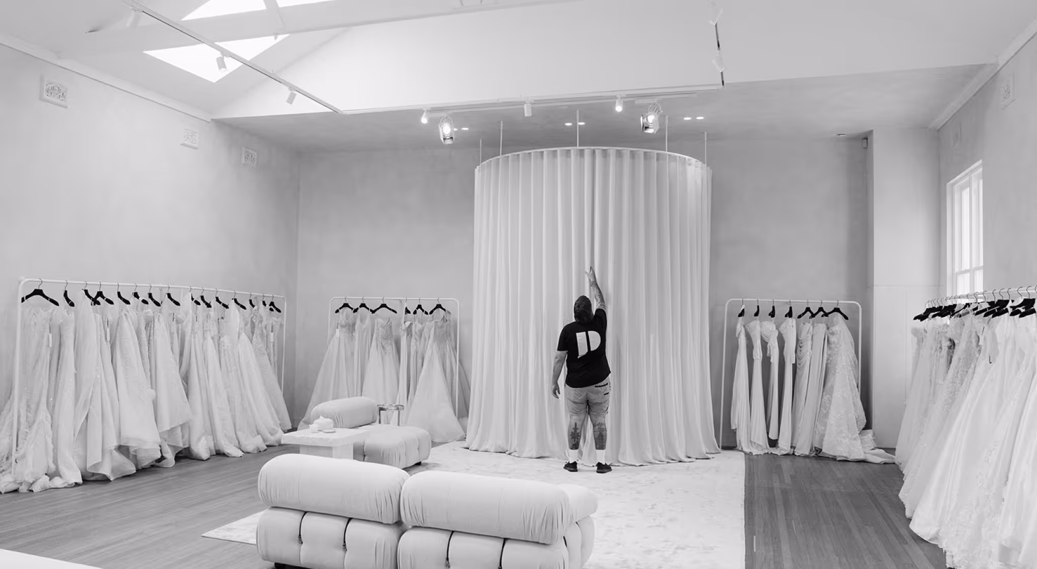 Man standing in bridal boutique with wedding dresses on racks surrounding a circular curtain.