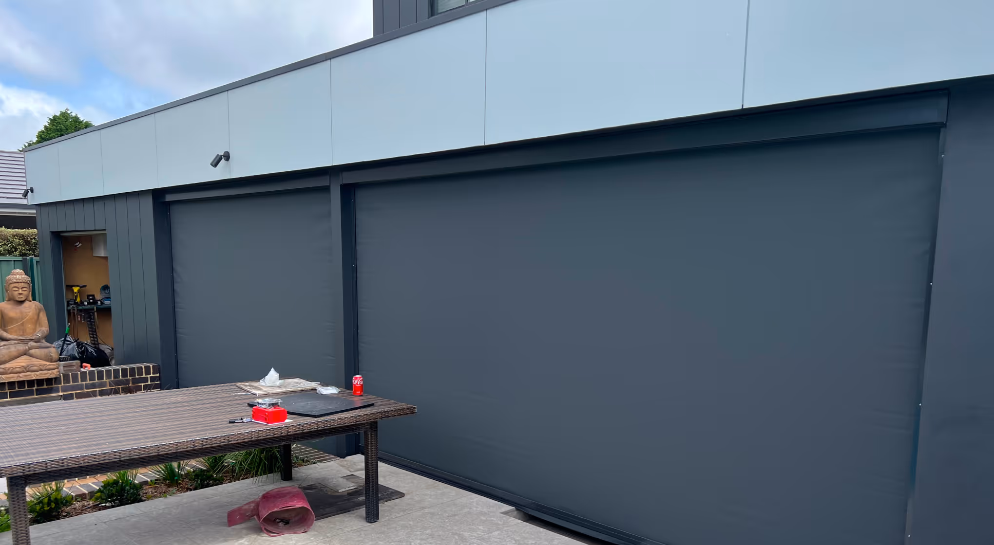 Outdoor patio with a large table, dark roller blinds on building, and a Buddha statue on a brick platform.