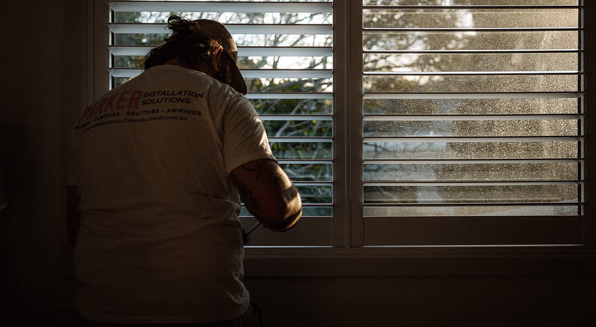 Person installing or adjusting white window shutters with sunlight and dust particles visible outside.
