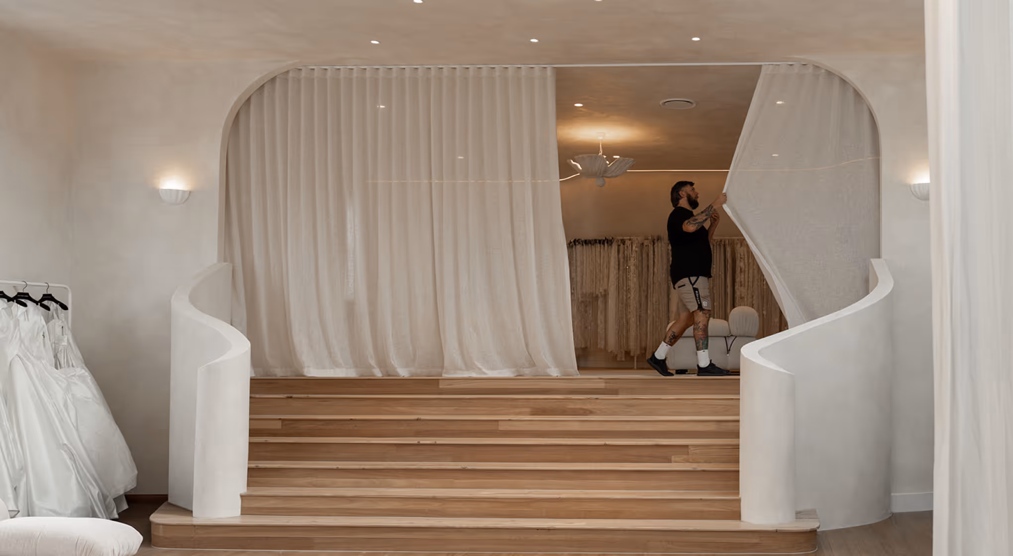 Man with tattoos draws back large white curtain in elegant minimal bridal boutique with wooden stairs.