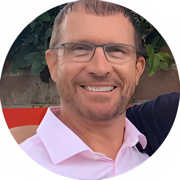 Smiling man with glasses, short hair, and a pink collared shirt outdoors near greenery and a wall.