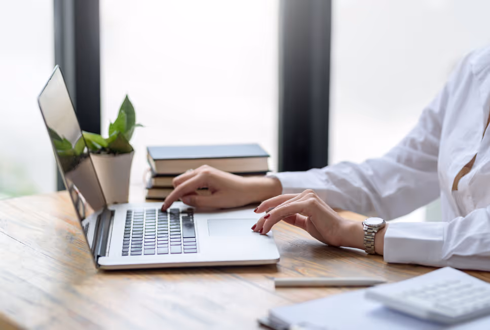 Person im weißen Hemd arbeitet am Laptop, eine Hand tippt auf der Tastatur, die andere liegt am Touchpad; Schreibtisch mit Pflanze, Büchern und Unterlagen im Hintergrund