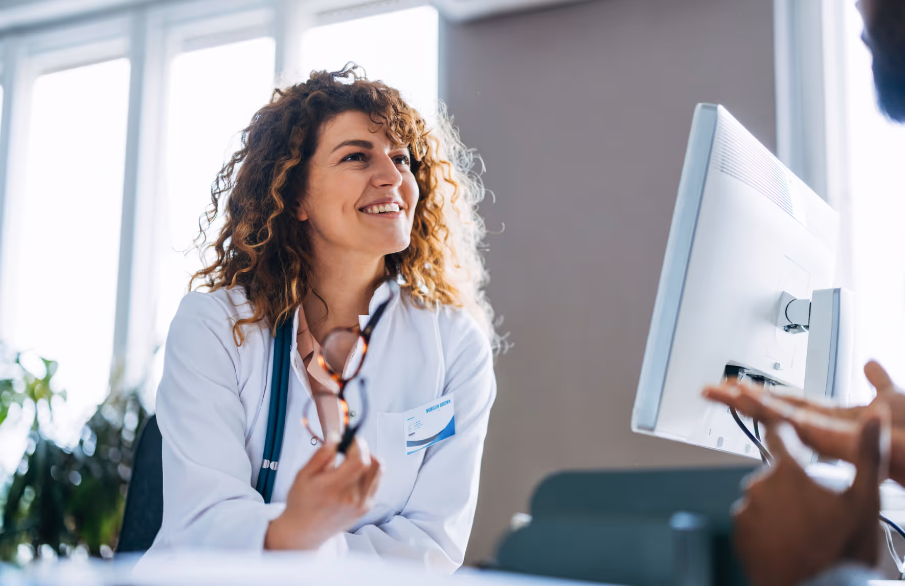 Lächelnde Ärztin im weißen Kittel sitzt am Computer, hält eine Brille in der Hand und spricht aufmerksam mit einem Patienten, dessen Hände erklärend gestikulieren