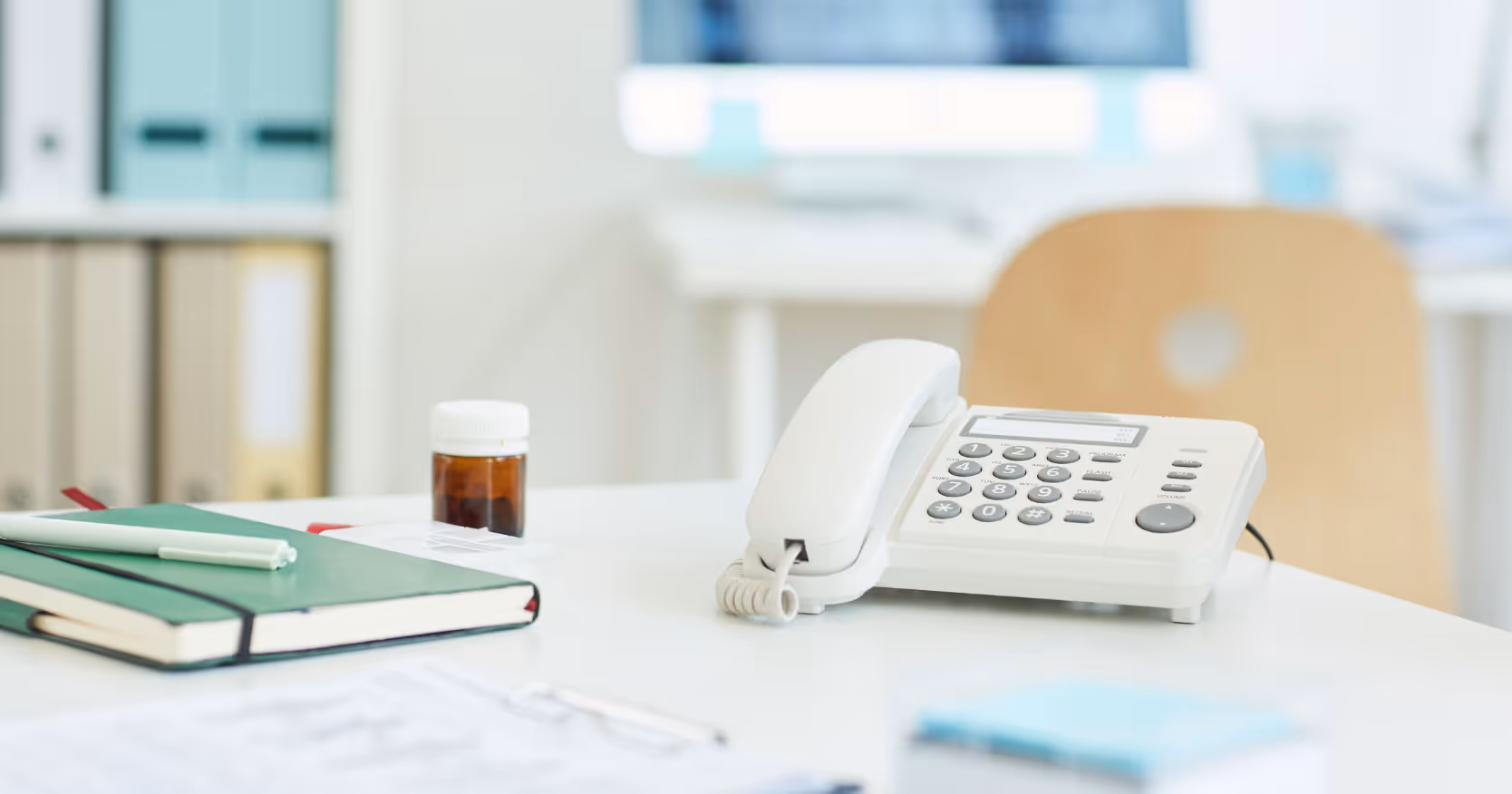 Weißes Telefon steht ruhig auf einem hellen Empfangstresen einer Arztpraxis, daneben liegen Notizbuch, Stift, Medikamentenbehälter und Unterlagen; die Szene vermittelt Entlastung, Ordnung und weniger Telefonstress am Praxisempfang.