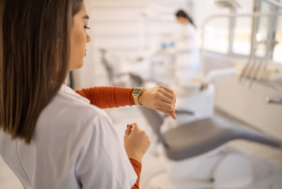 Mitarbeiterin in einer hellen Praxis schaut konzentriert auf ihre Smartwatch und prüft die Uhrzeit; im unscharfen Hintergrund ist medizinisches Personal im Behandlungsraum zu sehen, was ständige Erreichbarkeit und zeitnahe Patientenkommunikation symbolisiert.
