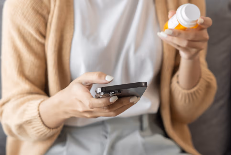 Person hält ein Smartphone in der einen Hand und ein Medikamentenfläschchen in der anderen; der Blick ist nicht sichtbar, die Gestik zeigt eine schnelle digitale Anfrage zu Medikamenten oder Rezepten über das Handy.