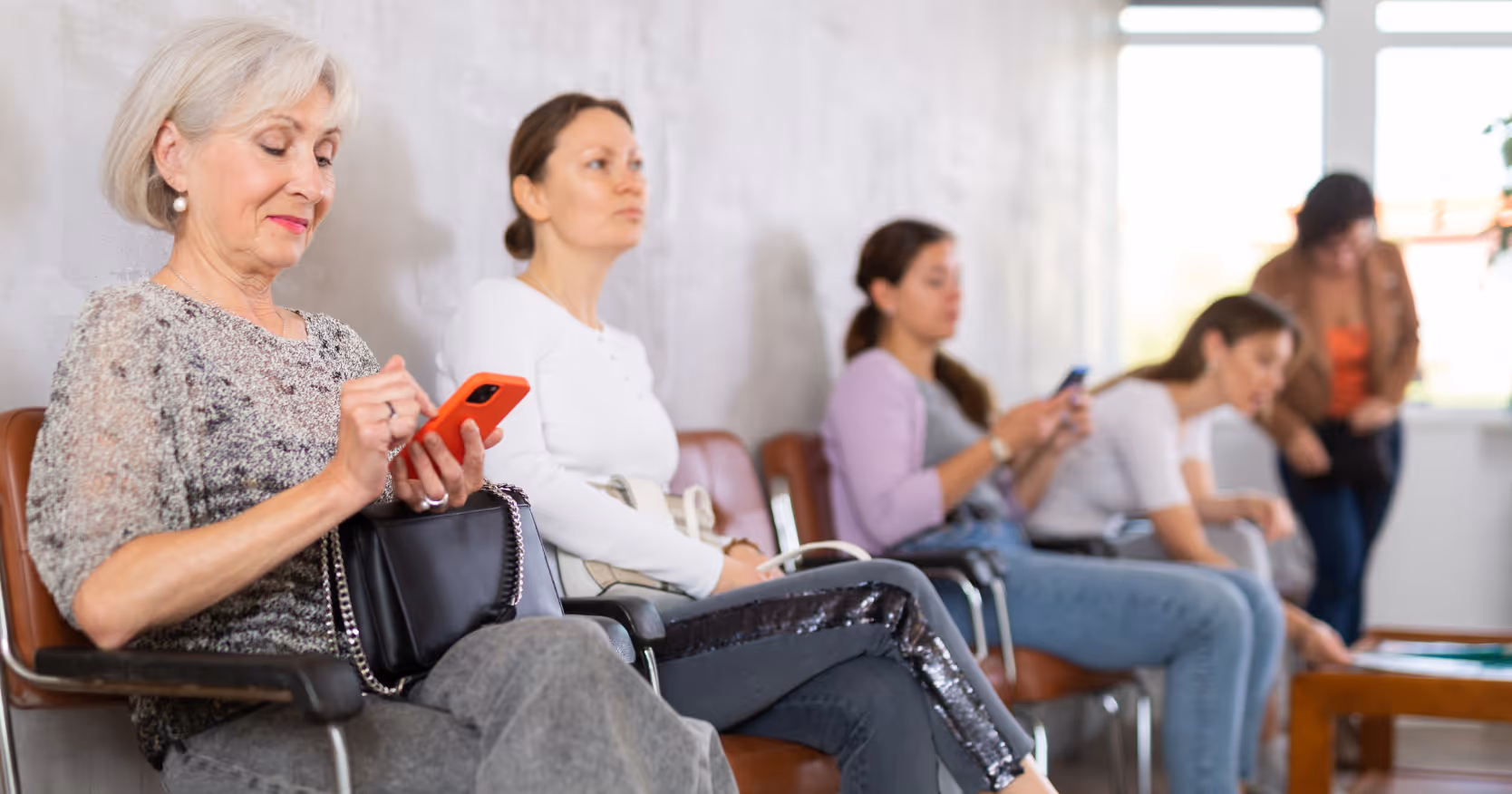 Mehrere Patientinnen sitzen im Wartezimmer einer Arztpraxis; eine ältere Frau im Vordergrund blickt konzentriert auf ihr Smartphone und nutzt es zur digitalen Patientenaufnahme.