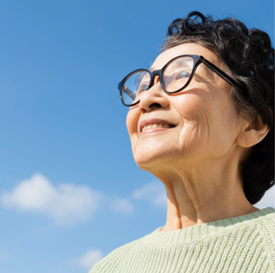 Smiling elderly woman wearing glasses and a green sweater looking up against a blue sky.
