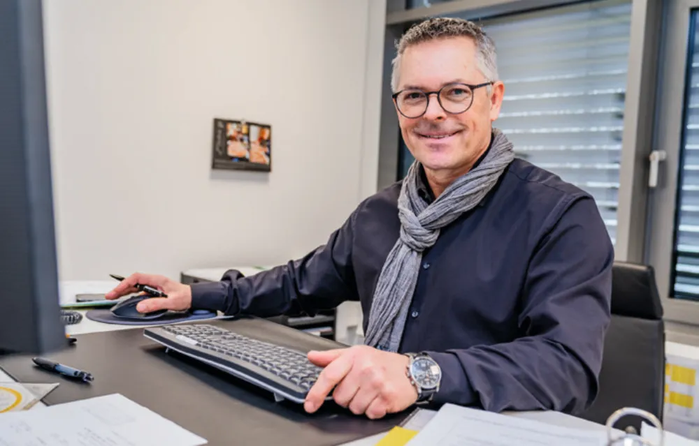 Mann mit Brille und Schal sitzt lächelnd an einem Schreibtisch mit Computer.