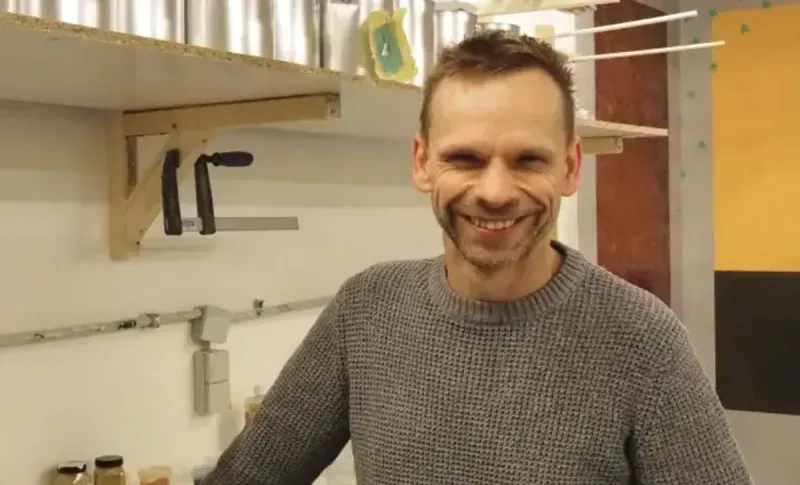 Ein lächelnder Mann mit grauem Pullover in einer Werkstatt oder Küche mit Regalen im Hintergrund.