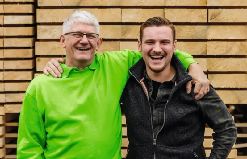 Zwei Männer, älter und jünger, lachen mit Armen umeinander vor Holzstapelwand.