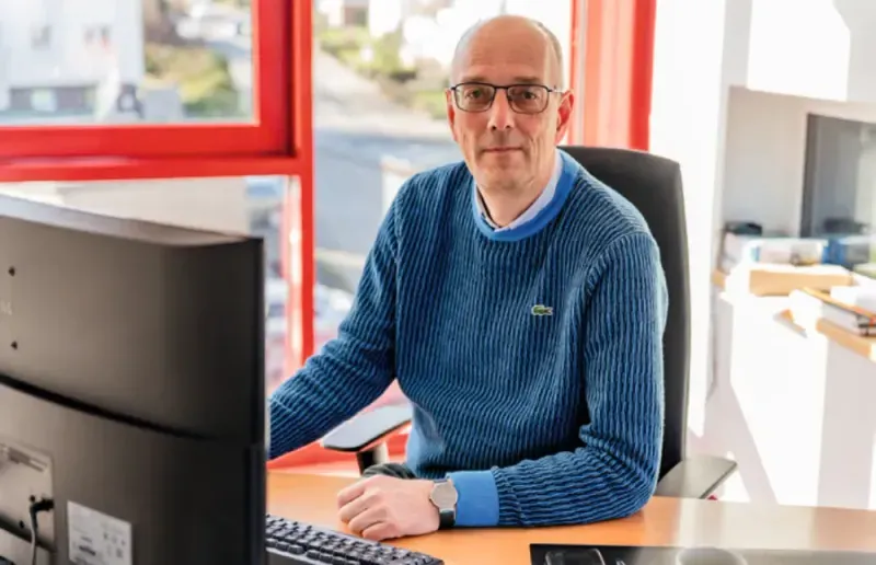 Älterer Mann mit Brille sitzt an Schreibtisch vor Computer in modernem Büro.