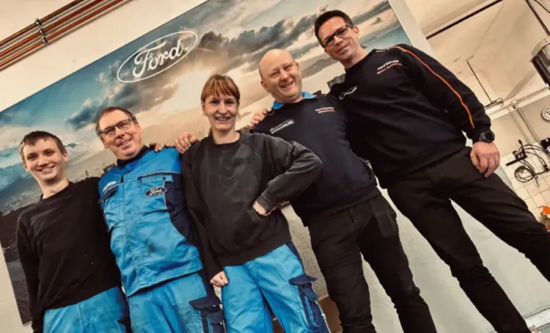 Fünf Mitarbeitende in Arbeitskleidung vor einem Ford-Logo auf einer Wand lächeln in die Kamera.