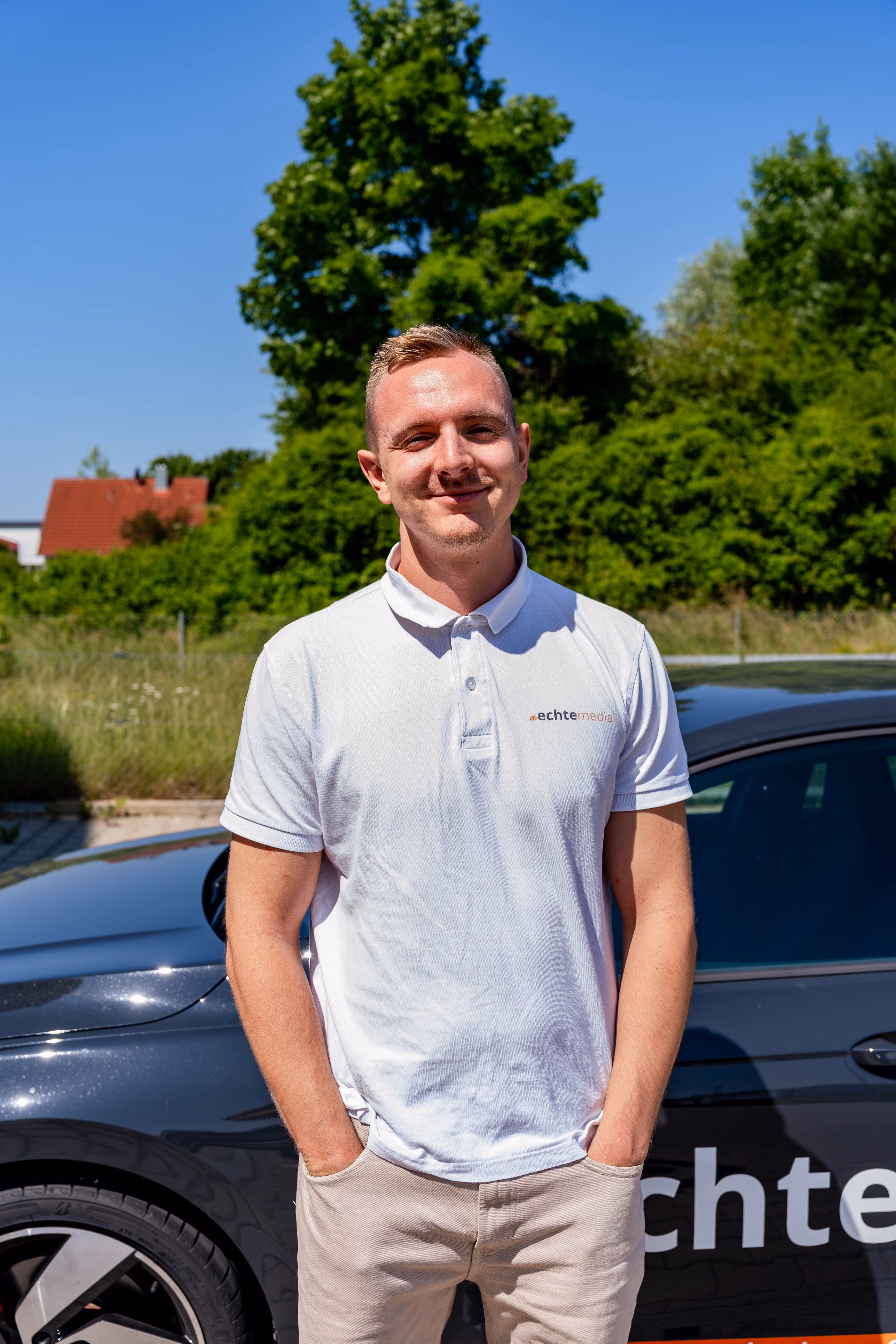 Junger Mann in weißem Polo mit echtemedia-Schriftzug steht vor schwarzem Auto im Freien.
