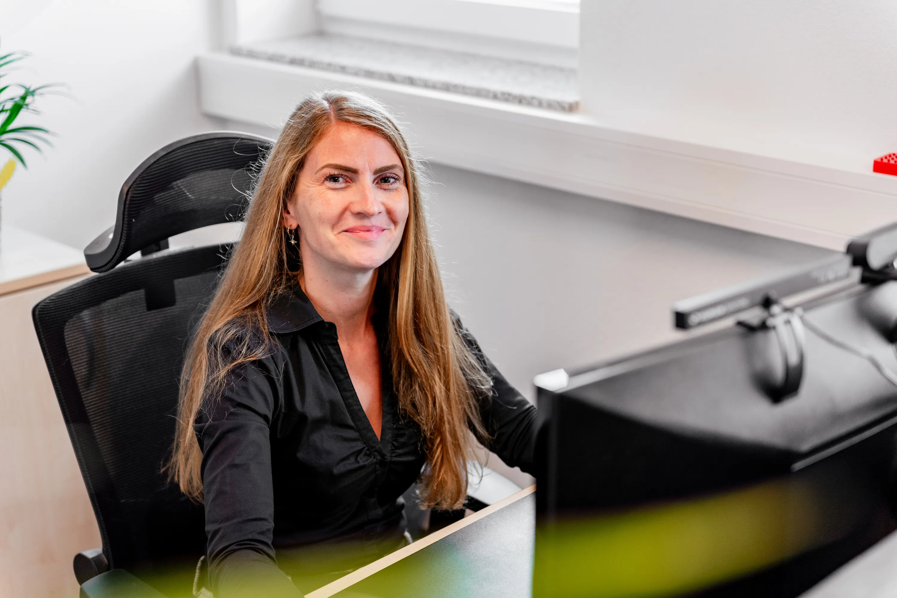 Frau mit langen blonden Haaren lächelt an ihrem Schreibtisch vor einem Computerbildschirm.