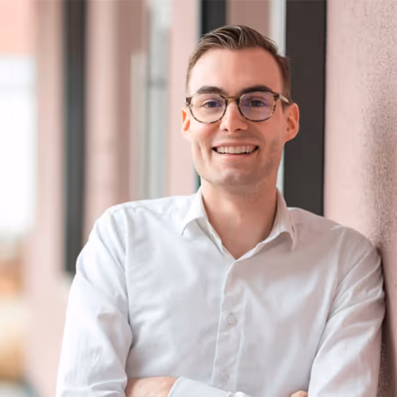 Lächelnder Mann mit Brille und weißem Hemd lehnt an Wand in hellem Büroflur.
