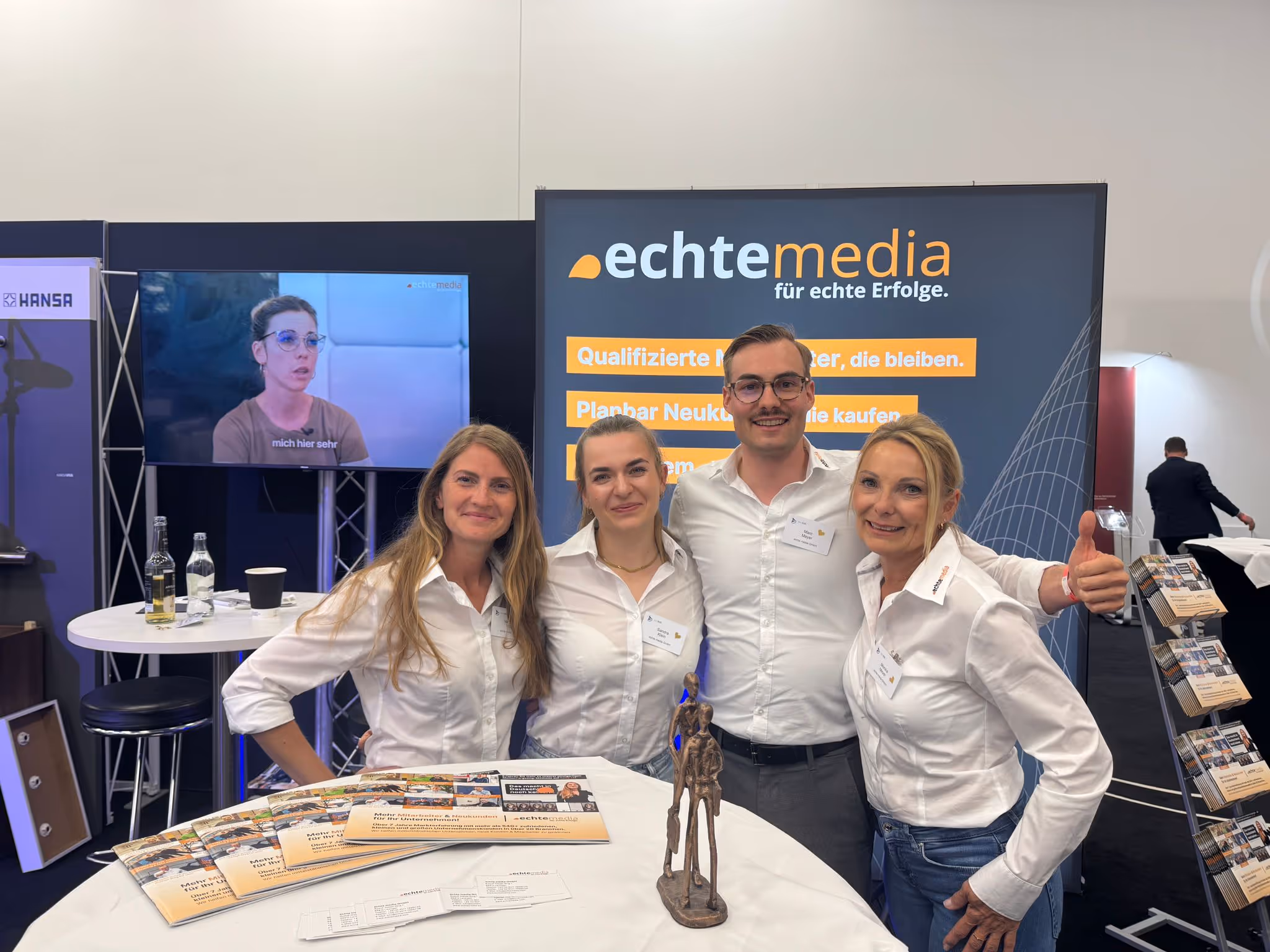 Vier Personen in weißen Hemden lächeln vor einem echtenmedia-Messestand mit Broschüren und Statue.