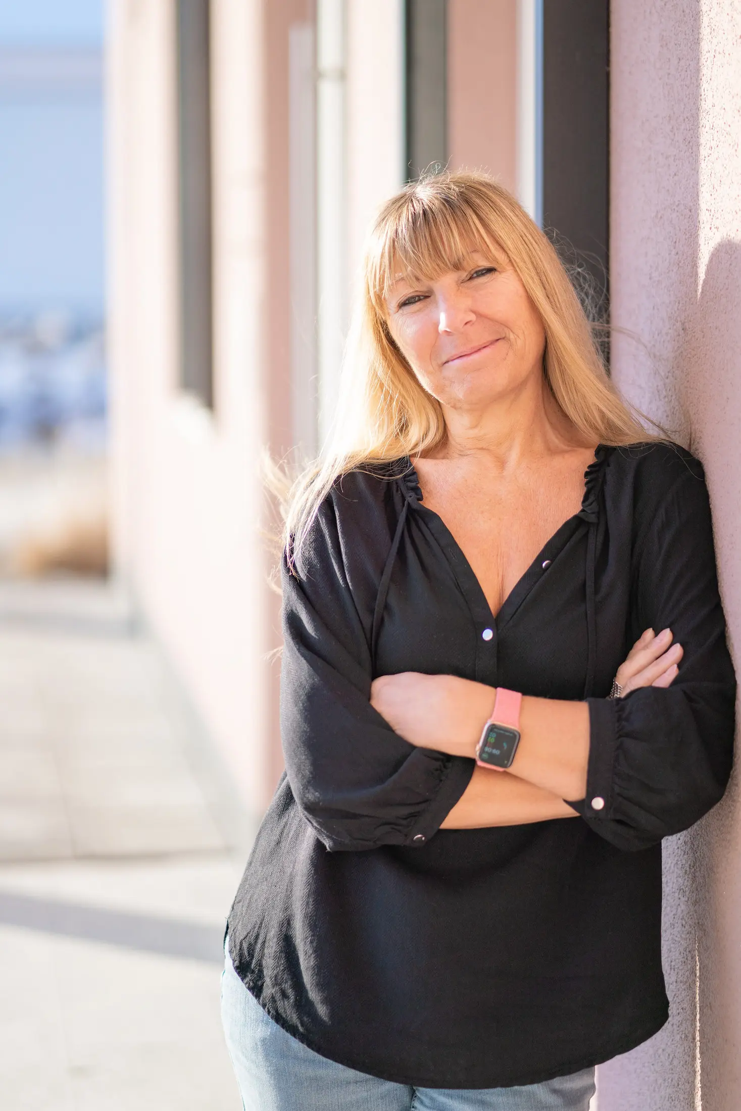 Frau mit langen blonden Haaren lehnt an Wand, trägt schwarze Bluse und rosa Smartwatch.