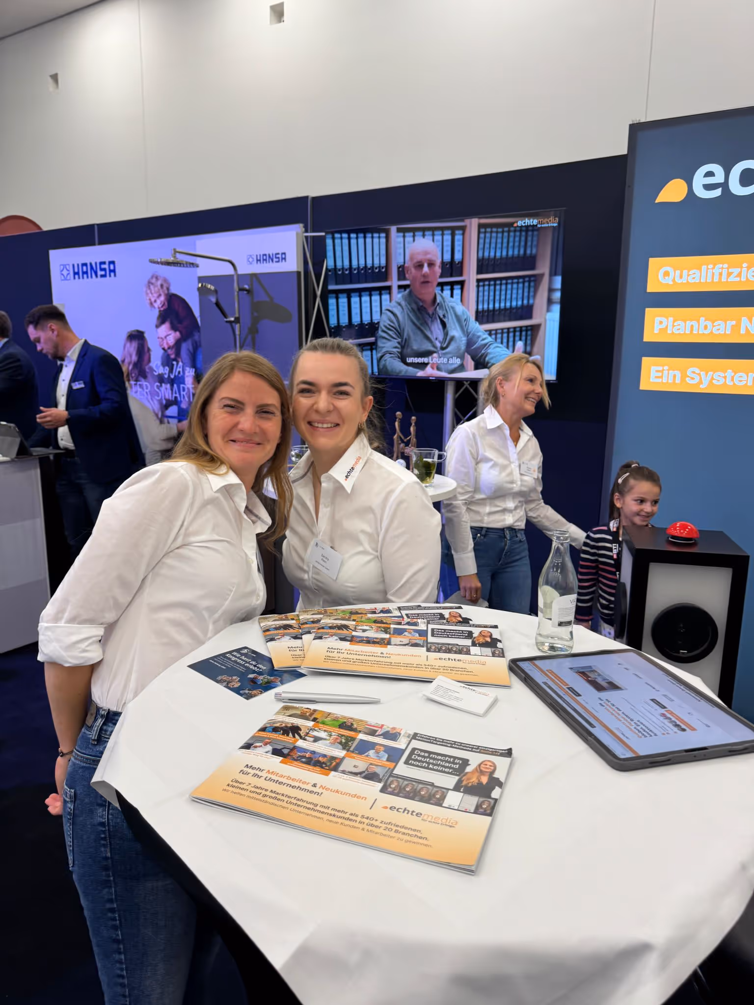 Zwei Frauen in weißen Hemden lächeln an einem Messestand mit Broschüren und Bildschirm im Hintergrund.