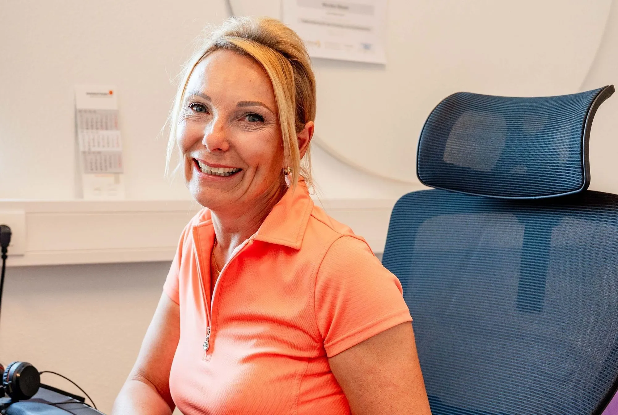 Lächelnde blonde Frau in orangem Polo sitzt an blauem Bürostuhl an einem Schreibtisch.