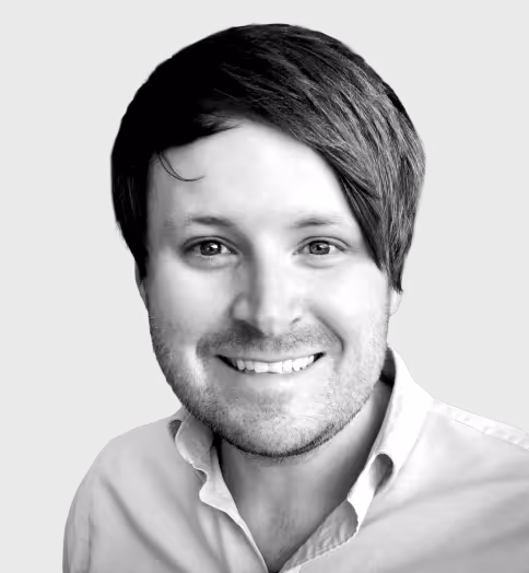 Smiling man with side-swept dark hair and light stubble, wearing a collared shirt against a plain background.