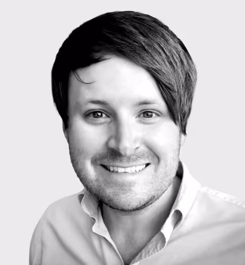 Smiling man with side-swept dark hair and light stubble, wearing a collared shirt against a plain background.