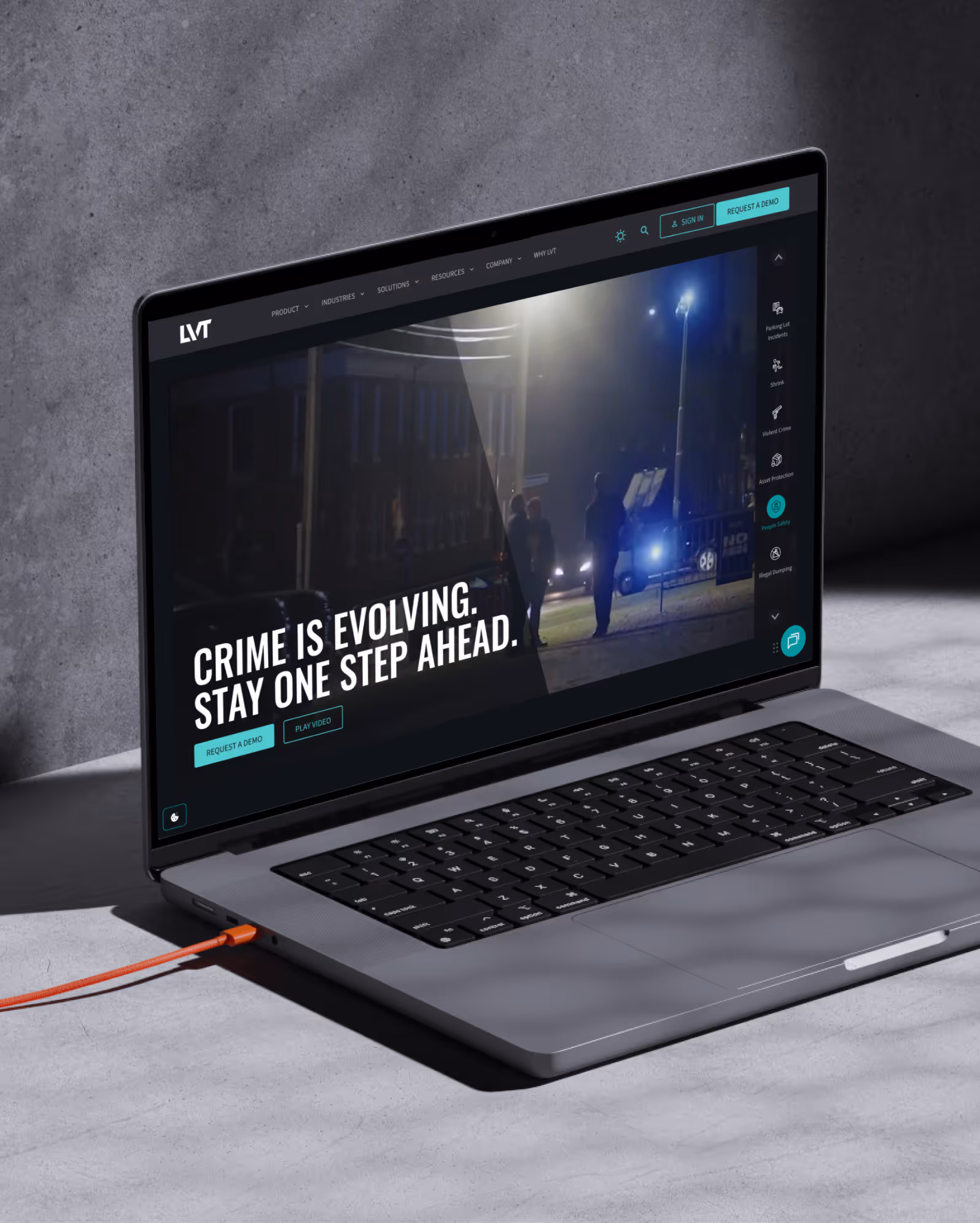 Laptop on a gray surface showing a security website with the headline 'CRIME IS EVOLVING. STAY ONE STEP AHEAD.' and nighttime surveillance footage.