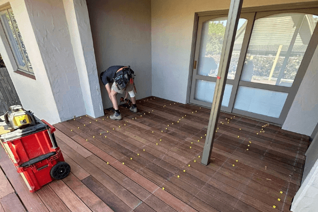 Worker installing wooden floor tiles on a porch with tools and spacers visible.
